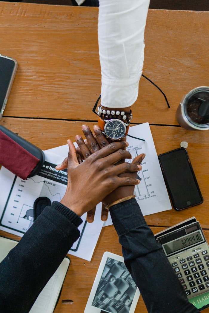 gallery-6 Top view of diverse hands stacked on office desk, symbolizing teamwork and collaboration.